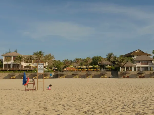 Sandy beach with thatched-roof villas and a shorebreak warning sign in Mui Ne