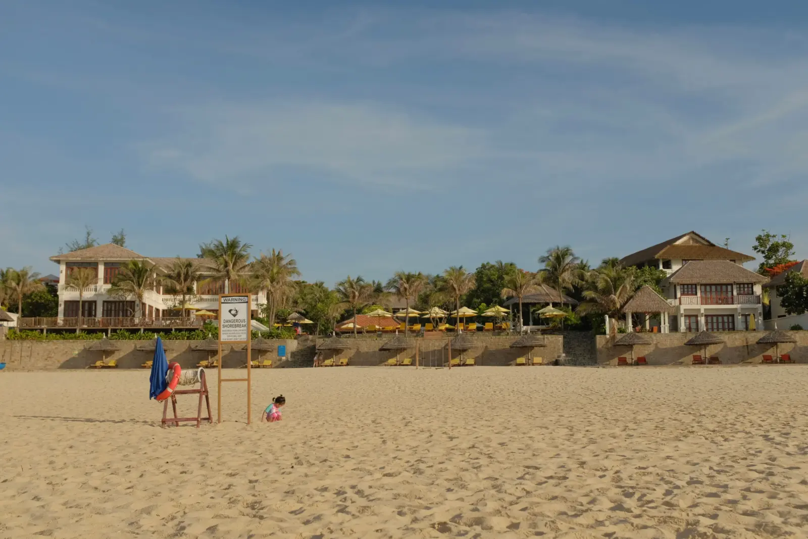 Sandy beach with thatched-roof villas and a shorebreak warning sign in Mui Ne