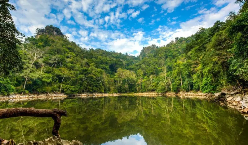 The serene, mirror-like surface of Ao Tien in Ba Be, surrounded by dense green forest and a bright, cloudy blue sky.