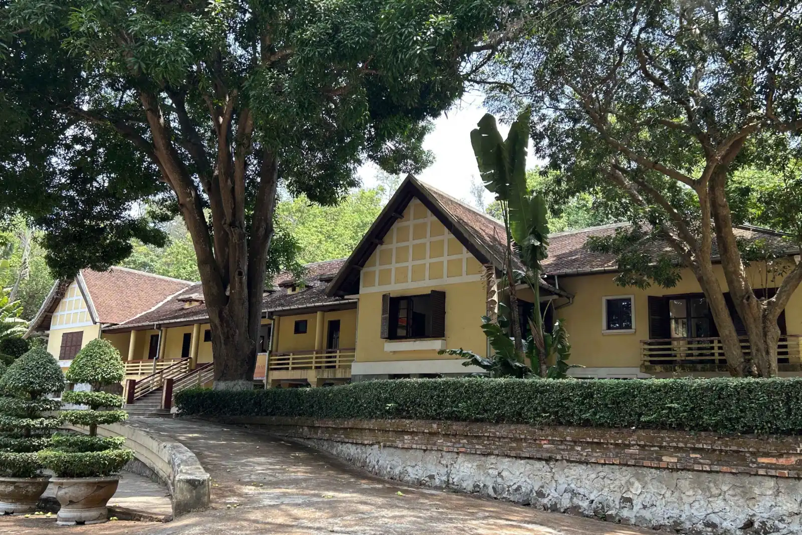 邦美蜀保大行宫（Bao Dai Palace）的黄色外墙，四周环绕着茂密的绿树和修剪整齐的灌木。